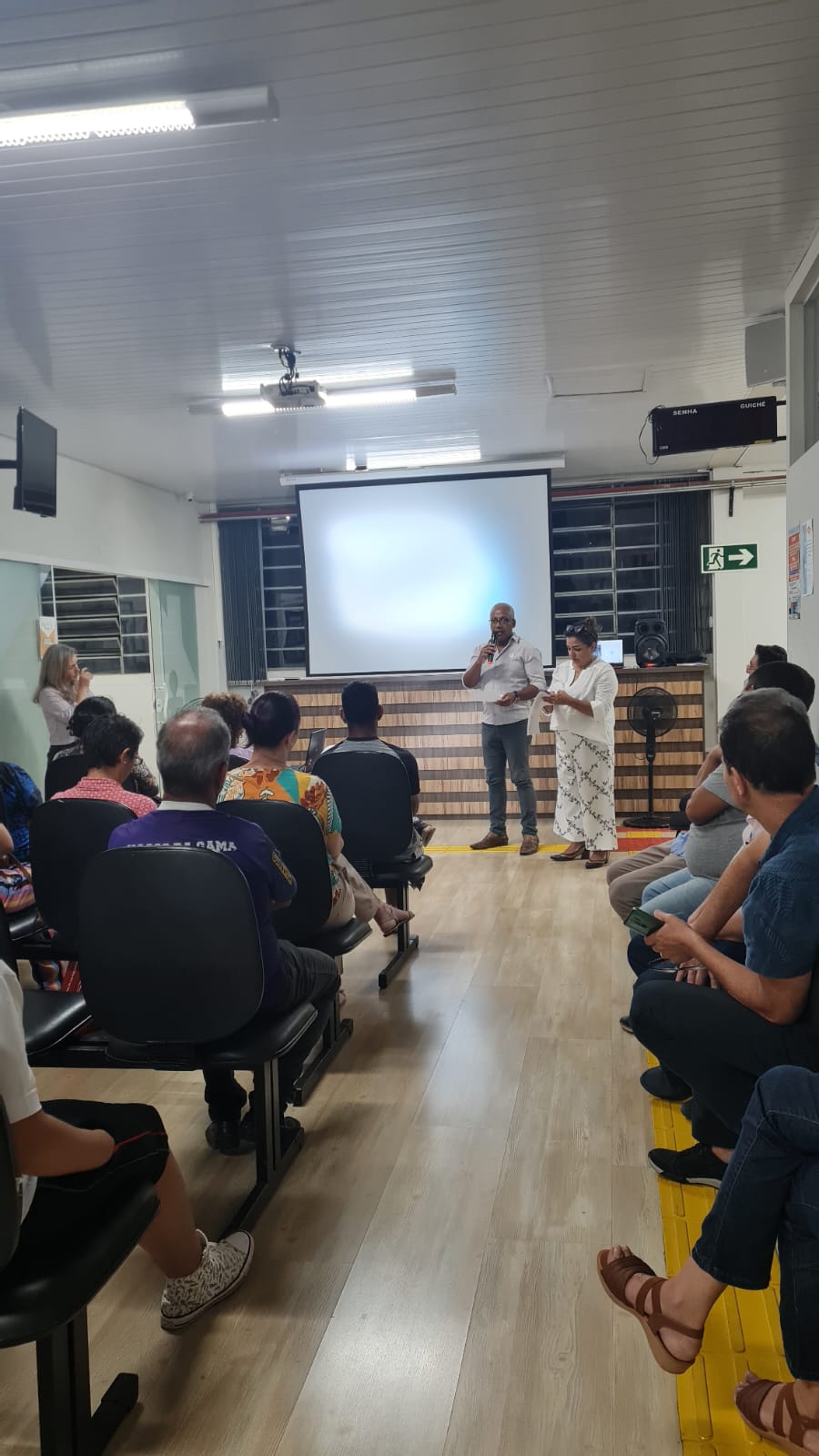 Descrição: Foto colorida. Foi fotografada com o público de costas para quem olha a imagem. Ao fundo da foto, de frente para o público está Denival Luiz de Oliveira, membro e advogado do Observatório da Inclusão e Gislaine do Rocio Rocha Simões da Silva, advogada e segunda tesoureira do Observatório, ambos em pé e um ao lado do outro, apresentam para o público presente, público este constituído das famílias dos associados, as demandas do Observatório da Inclusão.