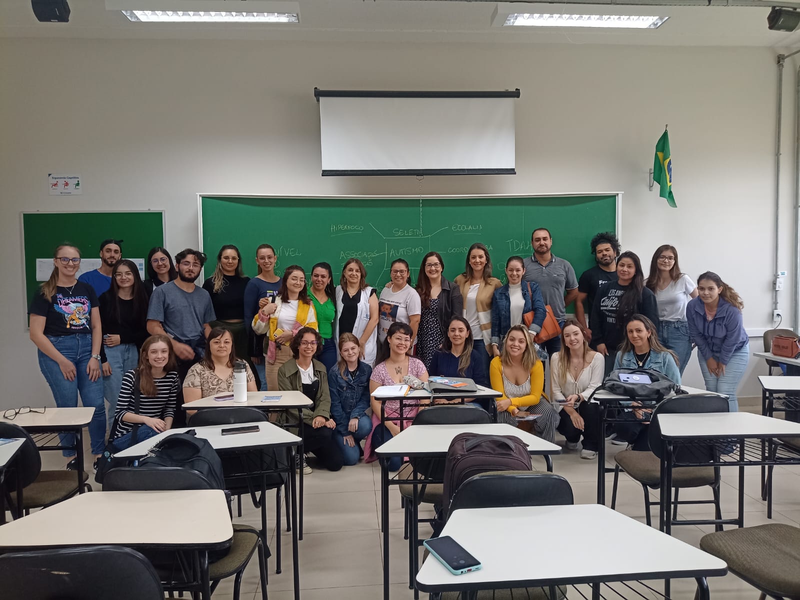 Descrição: Imagem fotográfica colorida em uma sala de aula. A imagem é composta no primeiro plano por várias carteiras, na cor branca com suas respectivas cadeiras na cor cinza, estão vazias, em seguida, após as carteiras, estão agachadas 9 jovens mulheres que estão sorrindo. Logo atrás delas em pé, também sorrindo, estão 19 pessoas. Sendo que destas pessoas, tem 4 homens. Ao fundo atrás das pessoas em pé, se vê um quadro escolar verde, ao centro da imagem. E ao fundo na lateral esquerda da imagem tem um quadro de recados também na cor verde. E na lateral direita da parede de cor branca tem a bandeira do Brasil.